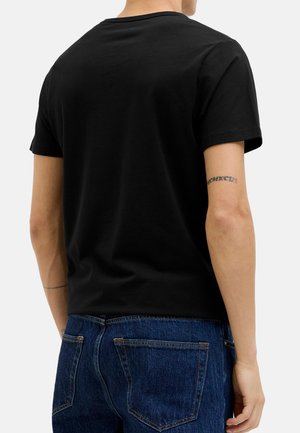 Persona che indossa una t-shirt nera a maniche corte semplice e jeans di denim blu, vista da dietro con un tatuaggio sul braccio sinistro.
