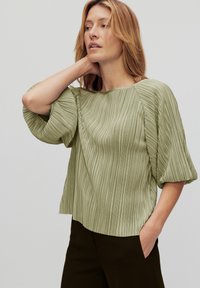 Vaaleanvihreä pleated-pusero, jossa on leveä kaula-aukko ja runsaat lyhyet hihat, yhdistettynä tummiin housuihin. Liukas tekstuuri, rento istuvuus.