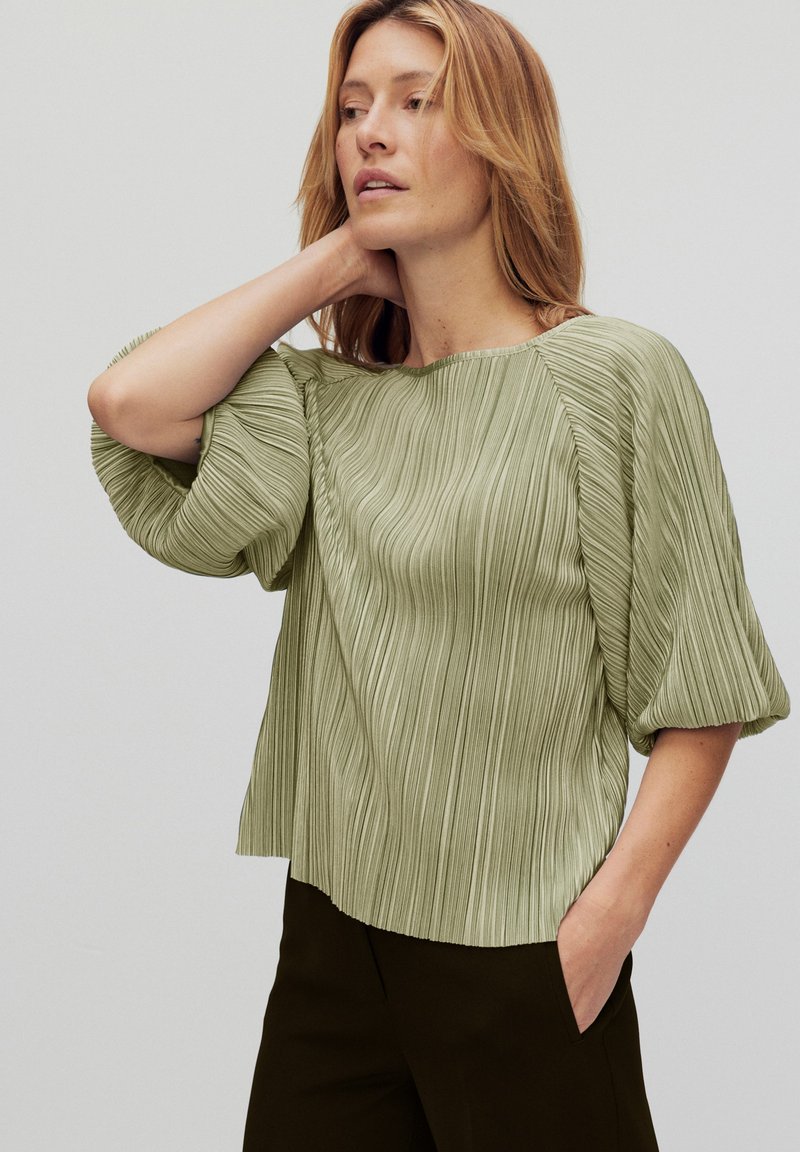 Vaaleanvihreä pleated-pusero, jossa on leveä kaula-aukko ja runsaat lyhyet hihat, yhdistettynä tummiin housuihin. Liukas tekstuuri, rento istuvuus.