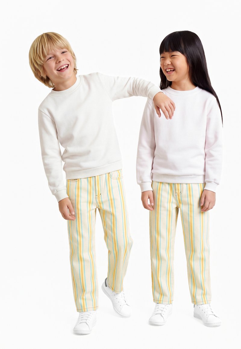 Deux enfants souriants debout, portant des sweats blancs, des pantalons rayés jaunes et bleus, et des baskets blanches sur un fond blanc uni.