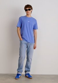 Svetlo modra bombažna majica z osrednjo logo, v kombinaciji s svetlo modrimi jeans hlačami in modrimi semiš supergami z belimi črtami, stoji na enobarvnem ozadju.