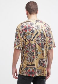 Kortärmad skjorta i beige med abstrakta geometriska mönster i rött, svart och blått. Har en avslappnad passform och en mjuk yta.