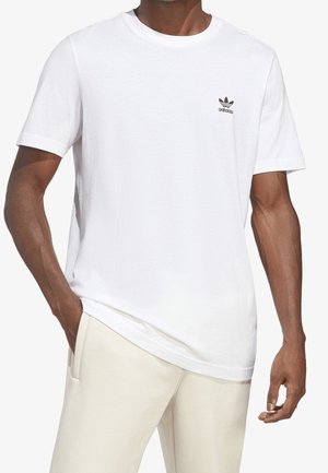 Homme portant un t-shirt blanc uni Adidas avec un petit logo noir sur la poitrine gauche et un pantalon beige clair, main dans la poche.