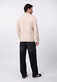 Vyras veidu į nugarą, dėvintis smėlio spalvos ilgomis rankovėmis marškinėlį, juodus džinsus ir juodus sportbačius su baltomis juostelėmis ant balto fono.