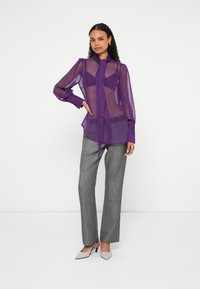 Paarse doorschijnende blouse met een striksluiting, lange mouwen en geplooide schouders, gecombineerd met grijze, op maat gemaakte broek en beige hoge hakken.