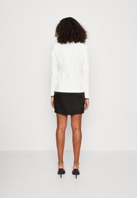 Veste blanche cintrée associée à une mini-jupe noire, présentant des textures lisses et une silhouette élégante. Des talons hauts noirs complètent la tenue.