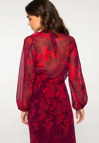 Robe transparente à motif floral rouge avec de longues manches bouffantes et un décolleté noué. Le tissu texturé crée un effet léger et superposé.