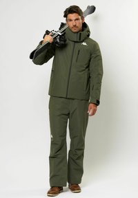 Olivazöld sídzseki és hozzáillő nadrág, mindkettő sima textúrával. A dzsekin magas gallér található, és jellegzetes logó van a mellkasán.