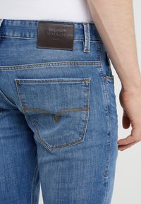 Blå denimjeans med en texturerad yta, med en läderetikett präglad med "JOOP!" och kontrasterande guldstygning på fickorna.