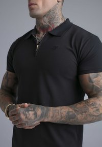 Persona musculosa con camiseta tipo polo negra con cremallera, con tatuajes coloridos extensos en el cuello, brazos y manos, de pie contra un fondo gris.