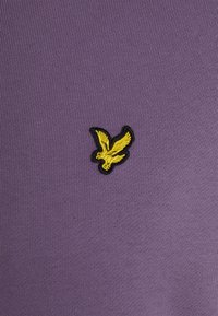 Tejido de punto púrpura con una superficie texturizada; presenta un logotipo de pájaro bordado en amarillo y negro en la parte frontal.