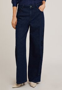 Jeans de mezclilla oscura de pierna ancha con intrincados bordados negros a lo largo del lateral. Presenta un diseño clásico de cinco bolsillos y un corte recto.