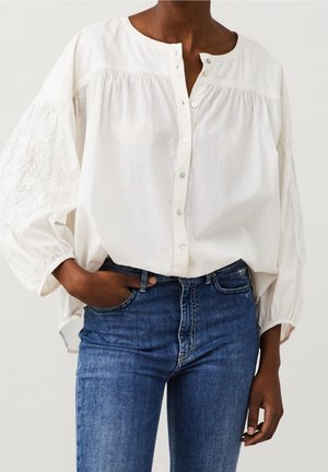 Personne portant un chemisier blanc ample avec des manches bouffantes brodées et un jean bleu taille haute, une main dans la poche, debout devant un fond uni.