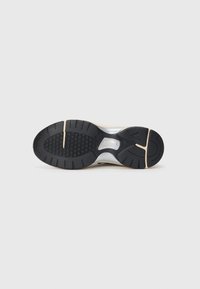 Suela de goma negra de una zapatilla beige que presenta un patrón de agarre hexagonal, ranuras de la suela y un puente de soporte en el mediopié en color plateado.