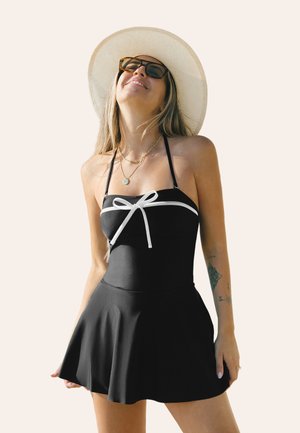 Zwarte bikini met een aansluitende top en een witte strikaccent, vloeiende rokontwerp en verstelbare bandjes. Textuurstof, minimale patronen.