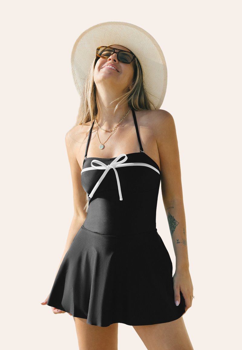 Maillot de bain noir avec un buste ajusté orné d'un accent en nœud blanc, design de jupe évasée et bretelles ajustables. Tissu texturé, motifs minimaux.
