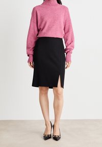 Pull en maille rose à col roulé avec épaules tombantes, associé à une jupe crayon noire dotée d'une fente sur le côté. Escarpins noirs à bout pointu avec accents dorés.