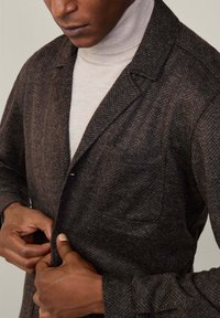 Cappotto marrone a spina di pesce con rever e tasca singola, indossato sopra un dolcevita chiaro. Tessuto strutturato, vestibilità su misura.