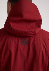 Rode waterproof jas met hoge kraag en capuchon. Voorzien van een zwart berglogo op de achterkant. Glad, gestructureerd stof met zichtbare naden.
