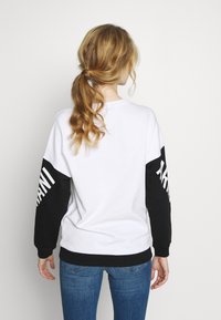 Weißer Pullover mit schwarzen Ärmeln, der ein großes Logo am linken Ärmel trägt, entworfen mit einer entspannten Passform und gerippten Bündchen.