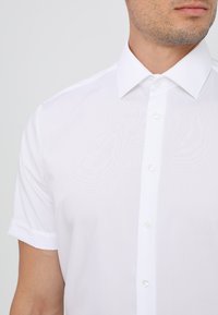 Camicia bianca a maniche corte con bottoni, realizzata in un tessuto liscio, con colletto classico e polsini arrotolati con dettagli dei bottoni a vista.