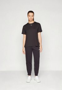 Mujer de pie con una camiseta negra de Puma, pantalones de chándal negros y zapatillas blancas frente a un fondo claro y liso.