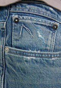 Modre kavbojke z poškodovanim videzom, s bakrenimi in črnimi kovinskimi dodatki ter zadnjim žepom z vidno šivalno nitjo in obrabljenimi detajli.