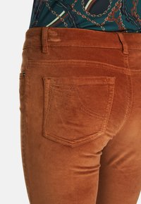 Braune Cordhosen mit samtiger Textur. Merkmale sind ein Fünf-Taschen-Design und eine Rückentasche mit dekorativem Stichdetail.