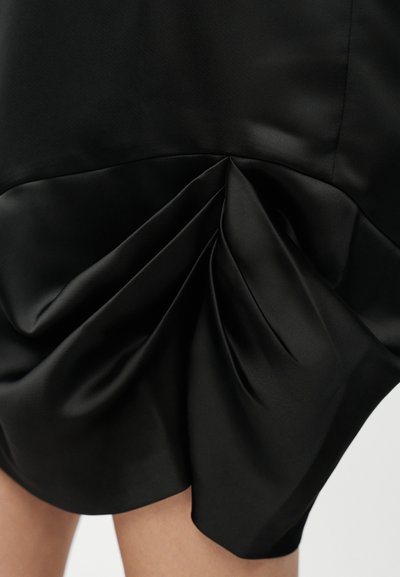 Φούστα από μαύρο σατέν ύφασμα με σούρες και draping πάνω από τα γόνατα, που αποκαλύπτει λεία και γυαλιστερή υφή με διακριτική λάμψη.