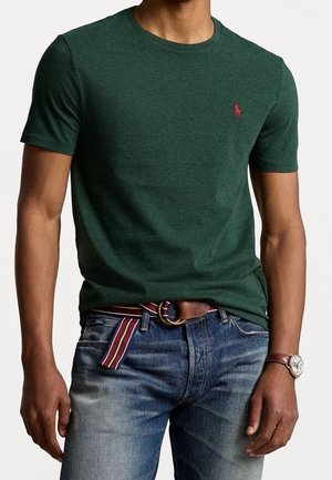 Homme portant un t-shirt vert foncé à manches courtes avec un petit logo rouge, un jean bleu, une ceinture en tissu rayée, une montre en cuir marron et une ceinture en cuir marron.