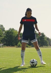Adidas sportkläder i marinblått med röda accenter; inkluderar en kortärmad topp och shorts. Spelaren står på gräs med en svart och vit fotboll.