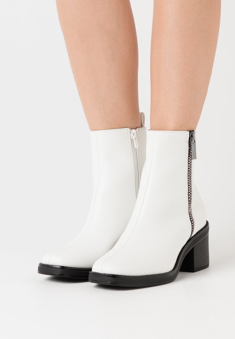 Bottes blanches en cuir mi-mollet avec talons noirs épais et fermetures éclair latérales portées sur des jambes nues, sur un fond blanc uni.
