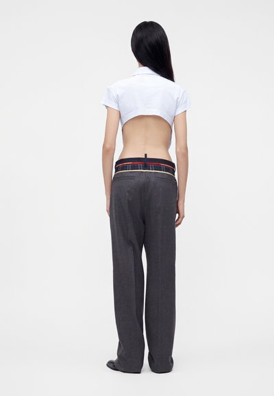 Femme aux longs cheveux noirs de dos portant un t-shirt blanc court, un pantalon large gris foncé taille haute et des chaussures noires.