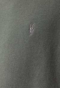 Sudadera de punto en verde oliva con un pequeño logotipo bordado de cráneo de cabra en gris claro. La tela tiene una textura suave y un diseño ajustado.
