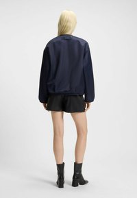 Navy bomber jacket with a smooth, shiny texture, featuring a relaxed fit and a ribbed collar, paired with black shorts and ankle boots.