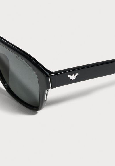 Gros plan sur des lunettes de soleil noires Emporio Armani, avec des verres foncés et un logo d’aigle argenté sur la branche.