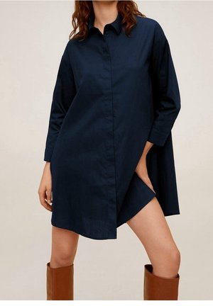 Femme portant une robe chemise bleu marine à manches longues avec une fente à l'avant, associée à de grandes bottes en cuir marron.