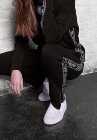 Chándal negro con rayas laterales blancas, con una tela suave y detalles de cremallera. El modelo lleva zapatillas blancas sobre una superficie oscura.