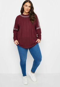 Burgundy långärmad topp med ribbad textur och rundad nederkant, med nitar längs ärmarna. Kombinerad med blå skinny jeans och vita sneakers.