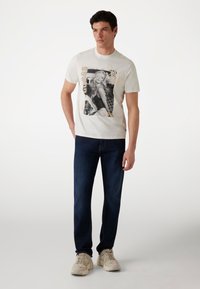 Balta medvilninė T-shirt su juodai baltu grafiniu raštu ir gėlių akcentais, kuriame užrašyta „GUESS“. Dėvima su tamsiai mėlynomis džinsomis ir smėlio spalvos sportbačiais.