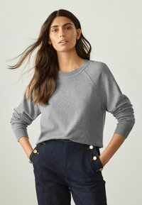 Grå sweatshirt med raglanärmar, ribbade manschetter och rundad halsringning, tillsammans med mörka byxor med guldknappdetaljer.