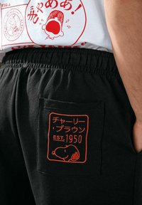 Black sweatpants feature an elastic waistband and a back pocket with orange embroidery displaying a cartoon dog and text in Japanese.