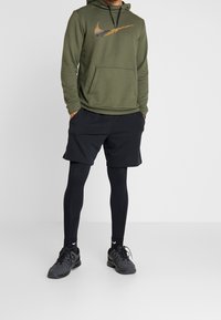 Sweat à capuche vert olive avec un logo graphique, short noir, leggings noirs et chaussures de sport noires. La tenue comprend une poche avant.