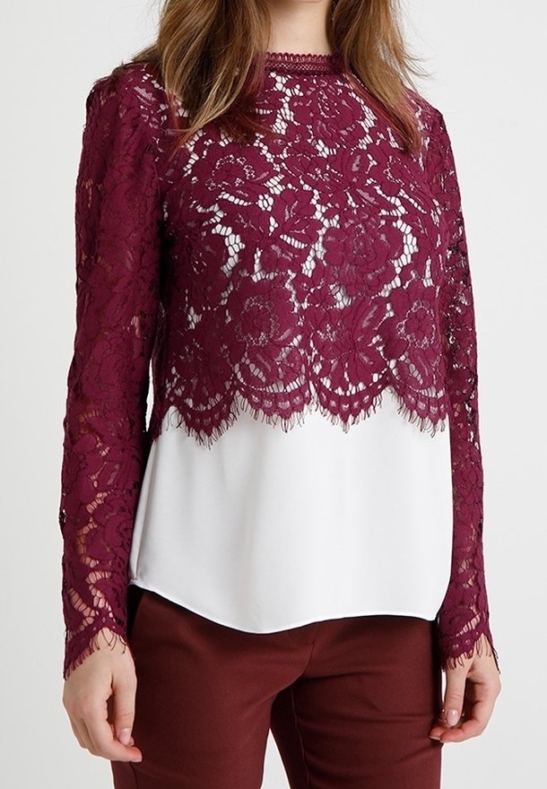 Femme portant un haut en dentelle bordeaux à manches longues par-dessus un chemisier blanc, assorti à un pantalon bordeaux, debout devant un fond uni.