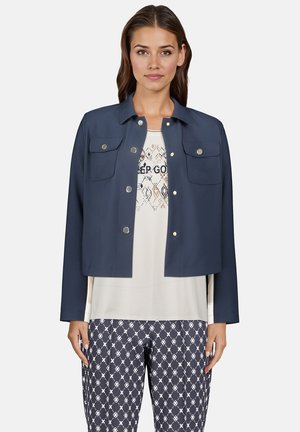 Marineblaue verkürzte Jacke mit Kragen, zwei Brusttaschen und Druckknopfverschlüssen. Über einem hellen Oberteil und gemusterten Hosen getragen. Glatte Stoffstruktur.