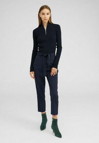 Marineblaues Rippenshirt mit langen Ärmeln und halbem Reißverschluss, kombiniert mit hoch taillierten marineblauen Hosen mit gebundenem Gürtel und grünen Wildleder-Stiefeletten.