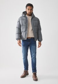 Hombre vistiendo una chaqueta acolchada gris, suéter beige, jeans azules y botas de cuero marrón, de pie contra un fondo blanco liso.