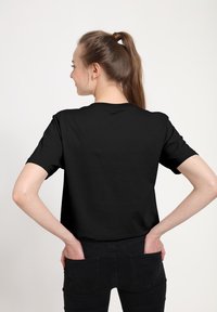 Černé tričko s krátkým rukávem a kulatým výstřihem, hladká tkanina, volný střih, nošeno s tmavými džíny, viděno zezadu.