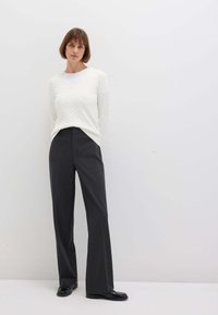 Donna in piedi contro un muro bianco, che indossa un maglione bianco a trecce, pantaloni grigi scuri a gamba larga e mocassini neri.