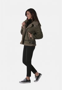 Veste en cuir retourné vert olive avec fermeture éclair, ceinture ajustable et deux poches avant, portée avec un jean noir et des baskets.